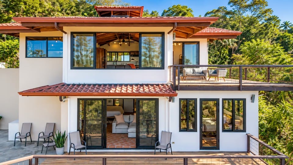 Two-story white home with red tile roofs, glass walls, an upper terrace, and a wood deck amid tropical forest.