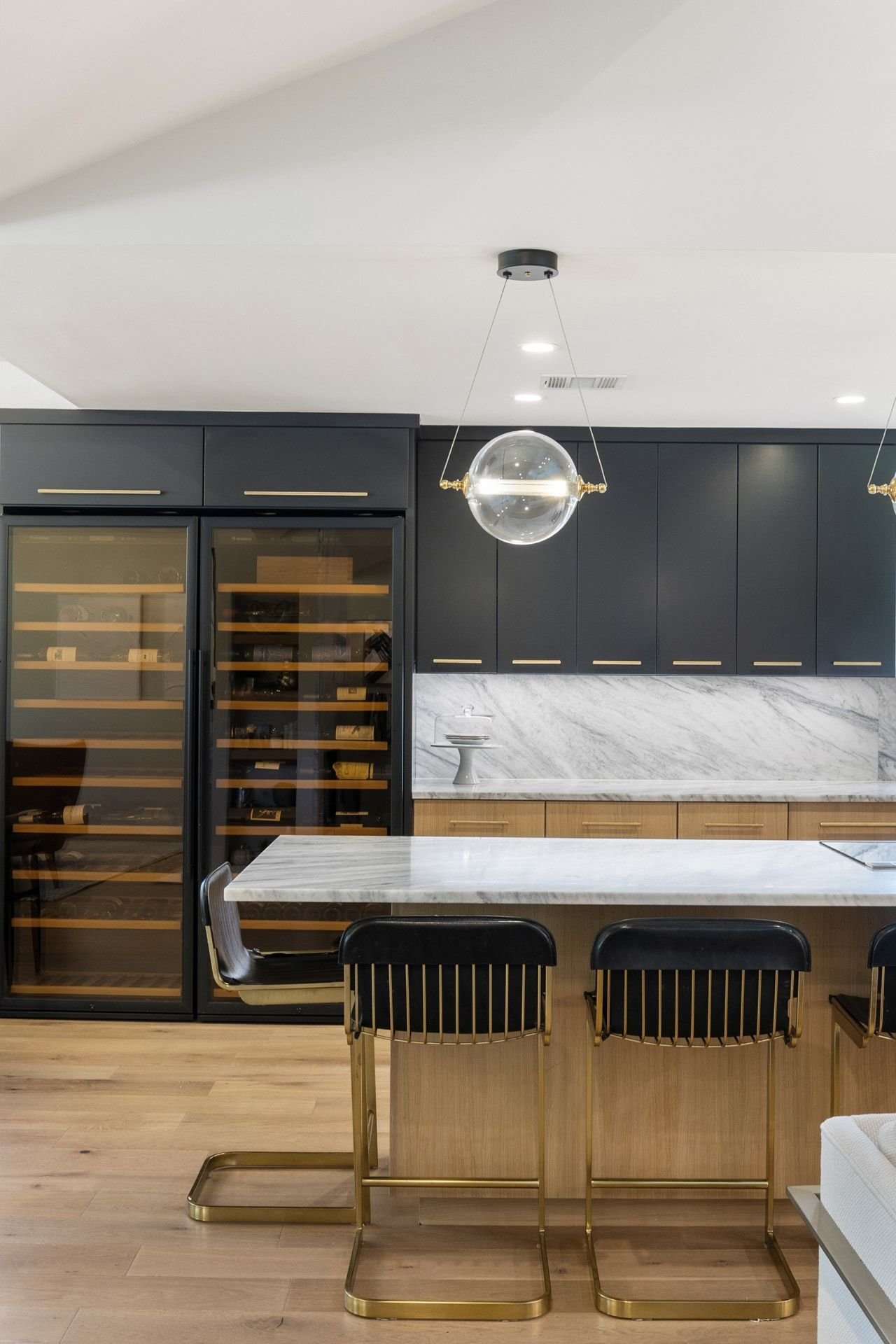 Modern kitchen with dark cabinets, glass-front wine refrigerators, marble island, and brass-accented stools.