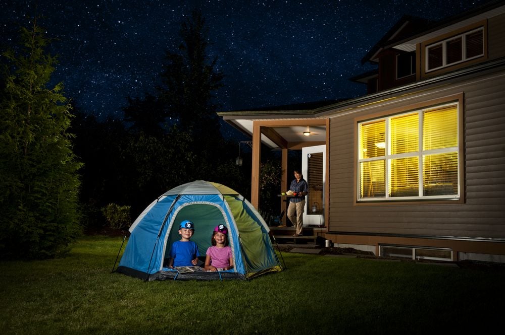 Two children sit in a backyard tent as an adult stands by the warmly lit house at night.