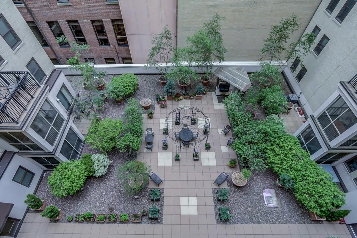 Apartment courtyard with a tiled patio, central metal table and chairs, and dense surrounding greenery.