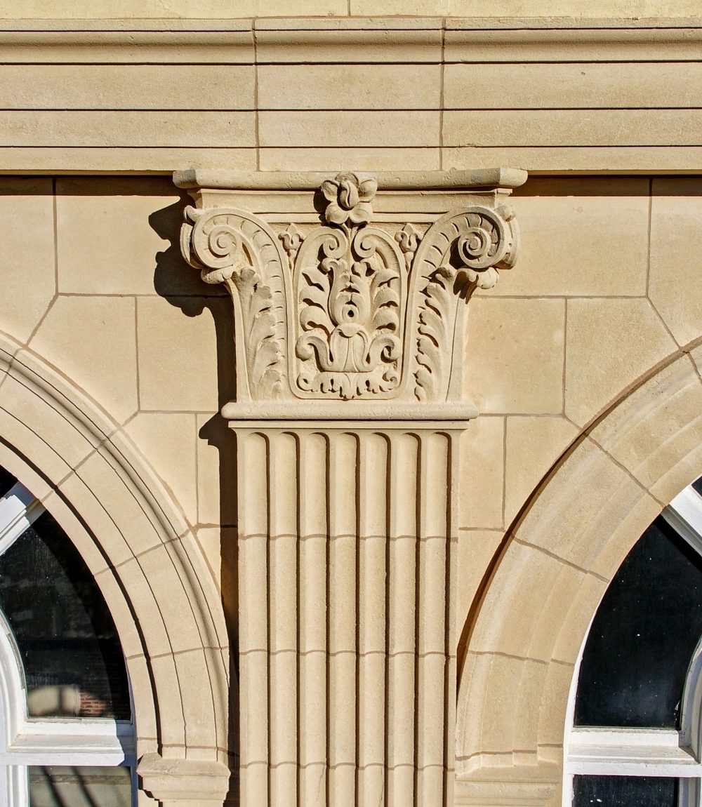 Carved stone capital and fluted column between arched windows on a building facade.