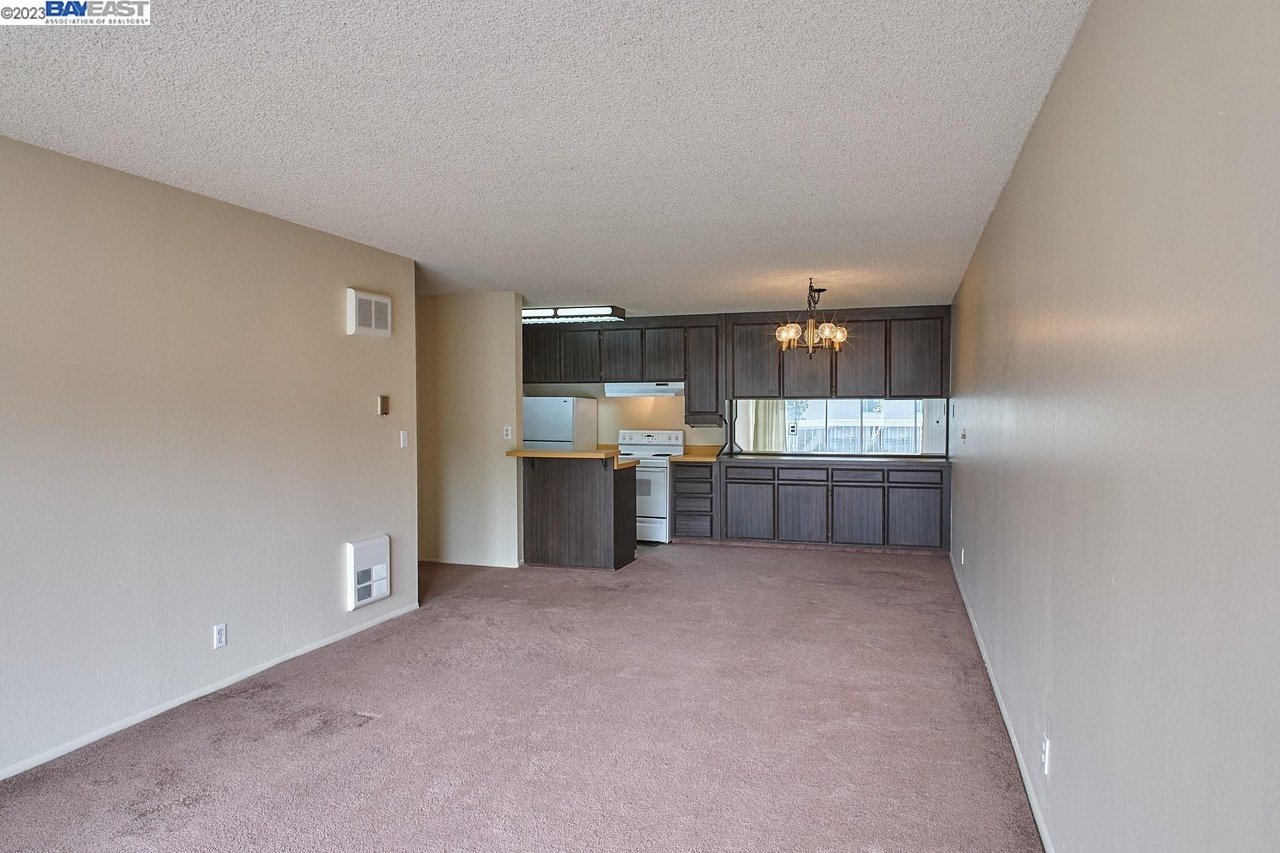 Empty living area with mauve carpeting and an open kitchen with dark cabinets and a peninsula.