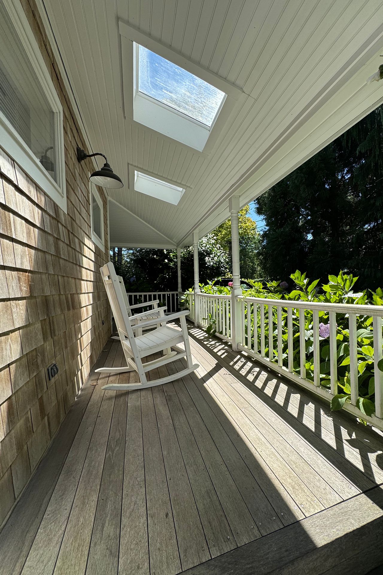 Covered porch with white rocking chairs, skylights, and leafy garden views.