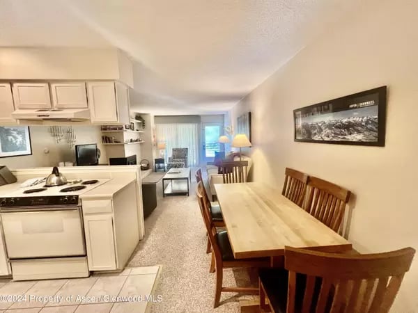 Compact home interior with white kitchen, wooden dining table, and living area at the back.