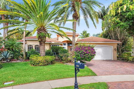 Single-story stucco home with a tile roof, palm trees, flowering shrubs, and a real estate sign by the sidewalk.