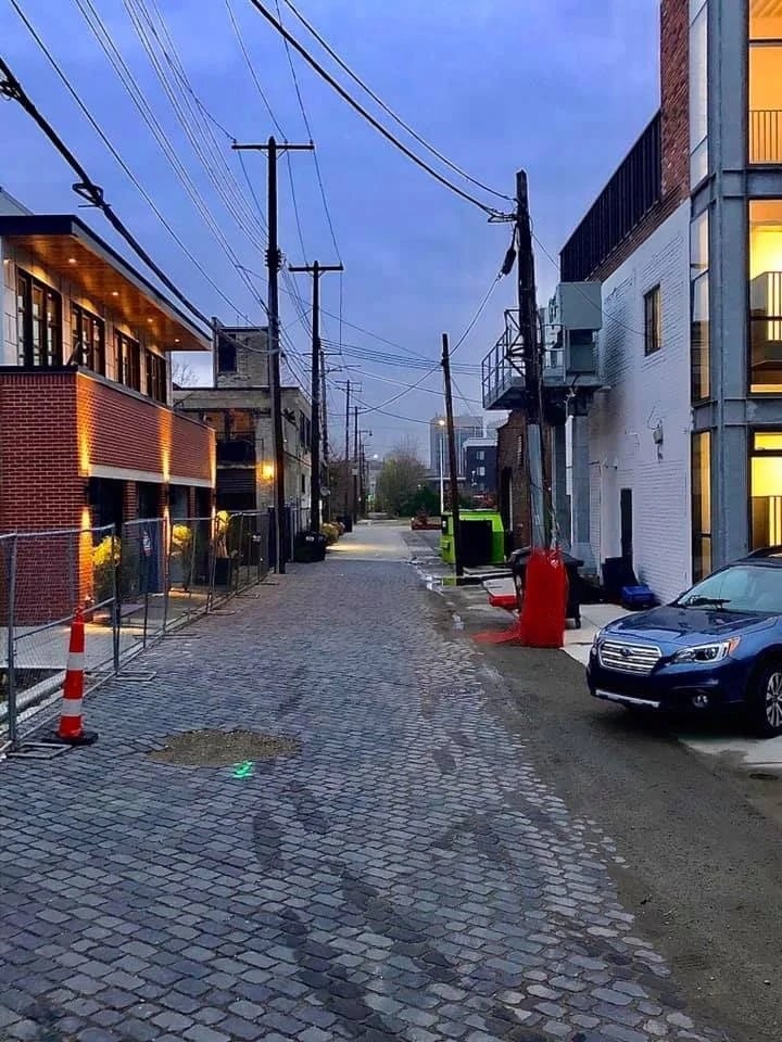 Cobblestone alley between brick and white mixed-use buildings at dusk, with utility wires overhead.