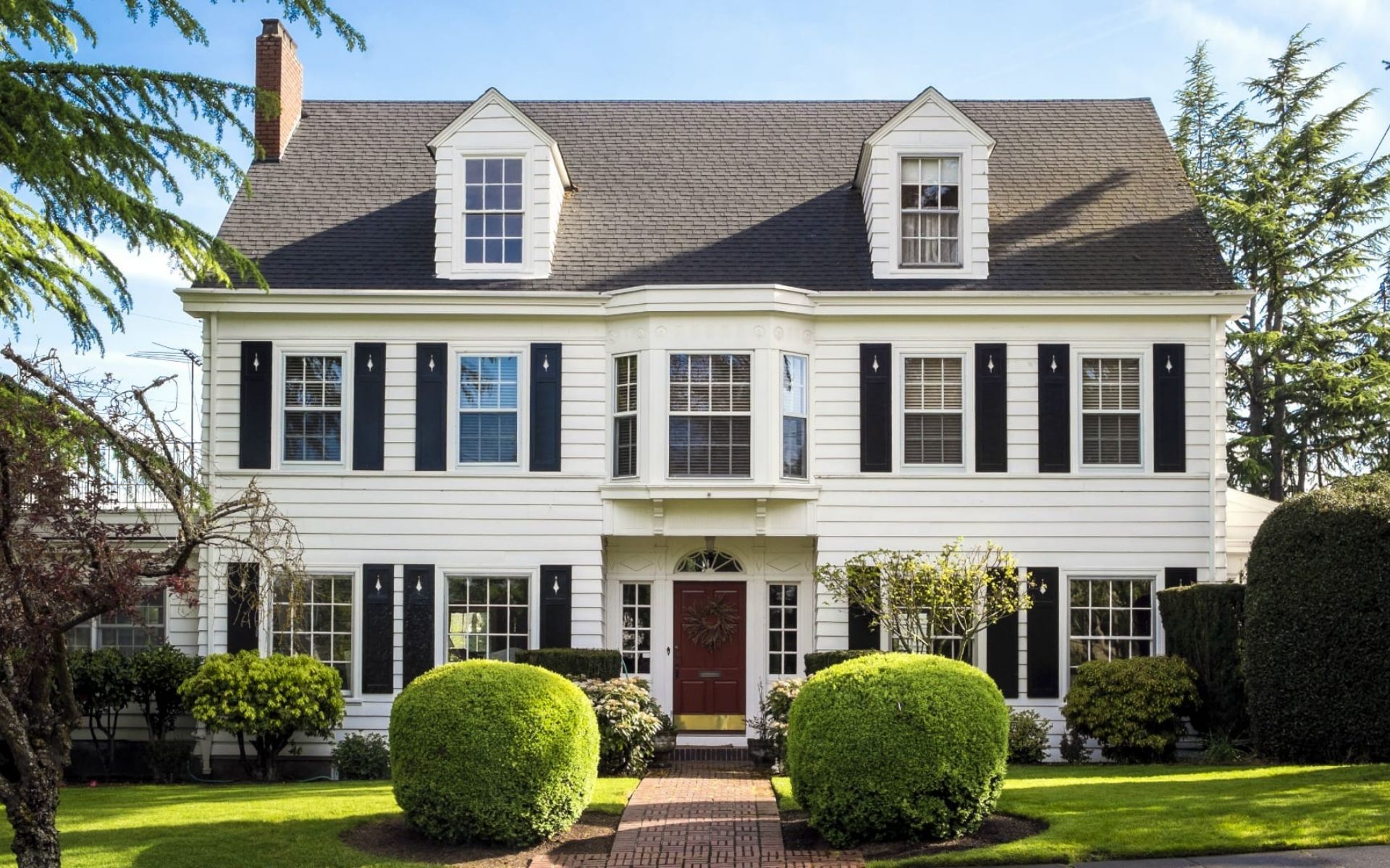White Colonial-style house with black shutters, dormers, a red door, and trimmed hedges.