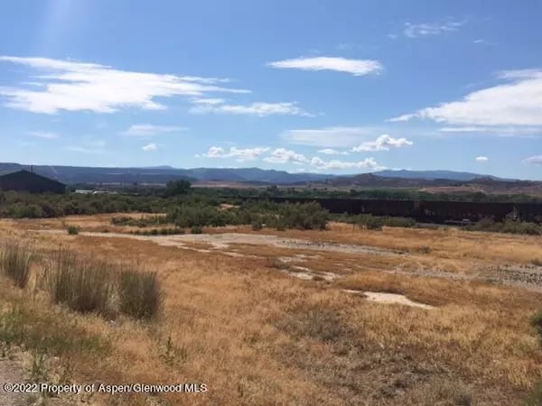 Dry grassland and scattered shrubs extend toward low hills beneath a blue, cloud-dotted sky.