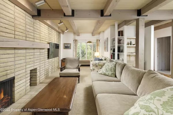 Living room with a cream sectional, pale brick fireplace, exposed beams, and windows overlooking trees.