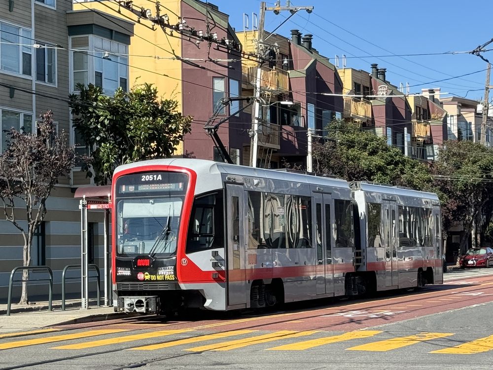 A J-Church Muni Metro light rail vehicle in San Francisco, traveling along Church Street with passengers visible through the windows. The surrounding area features residential buildings and tree-lined sidewalks, illustrating the integration of public transit within the neighborhood.