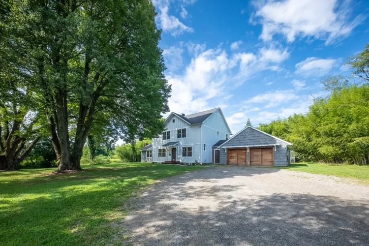 Two-story light-colored house with a large garage, gravel driveway, and mature trees.