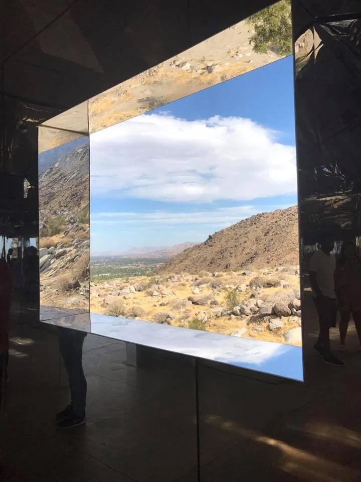 Mirrored art installation framing a rocky desert hillside, distant valley, and blue sky.