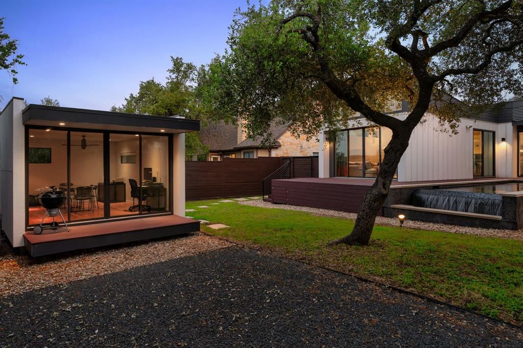 Modern home exterior with glass doors, timber deck, and a large oak tree at dusk.