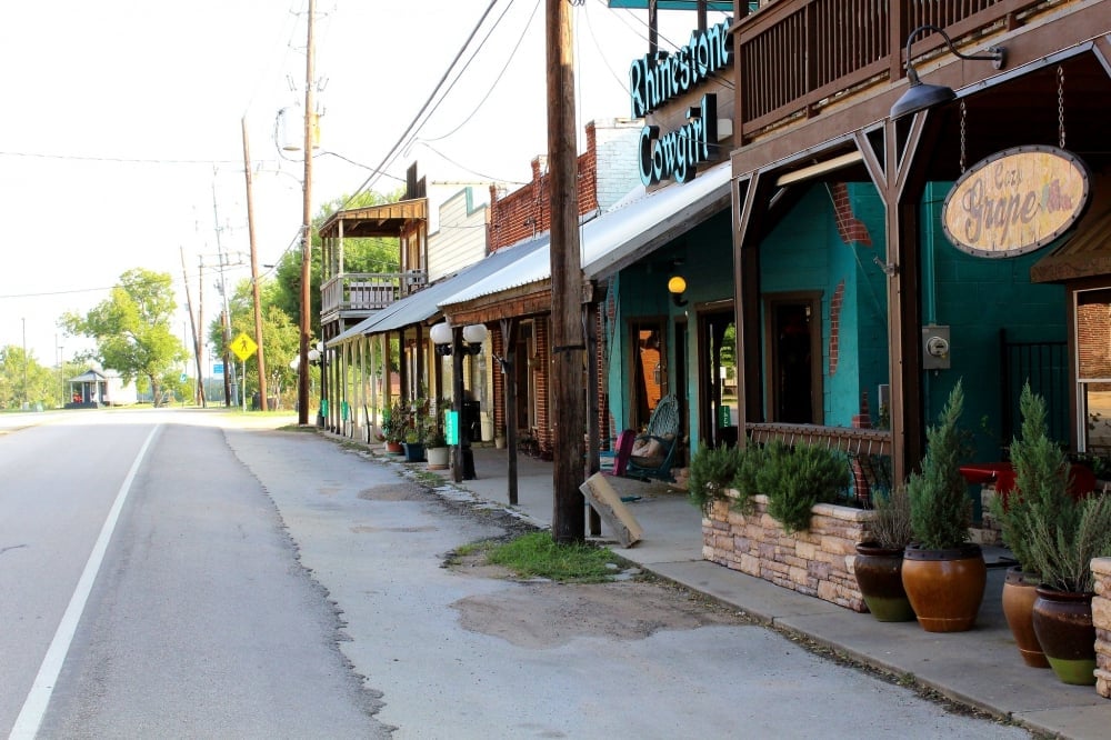 Small-town street lined with storefronts, including a turquoise shop with a wooden porch and potted plants.