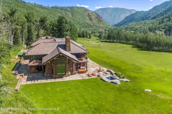 Timber mountain home with a stone patio in a broad green meadow beneath forested peaks.