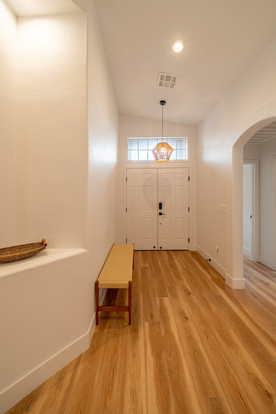 Entry foyer with a white double door, woven pendant light, narrow bench, and warm wood flooring.