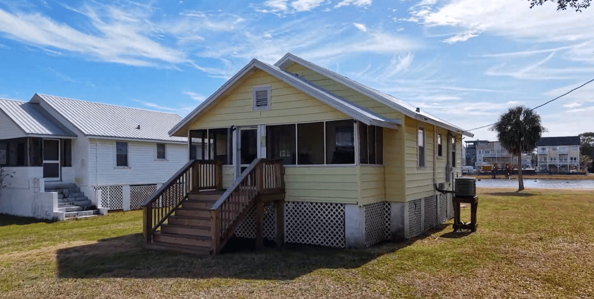 Raised yellow cottage with a screened porch and wooden stairs on a grassy lot beside the water.