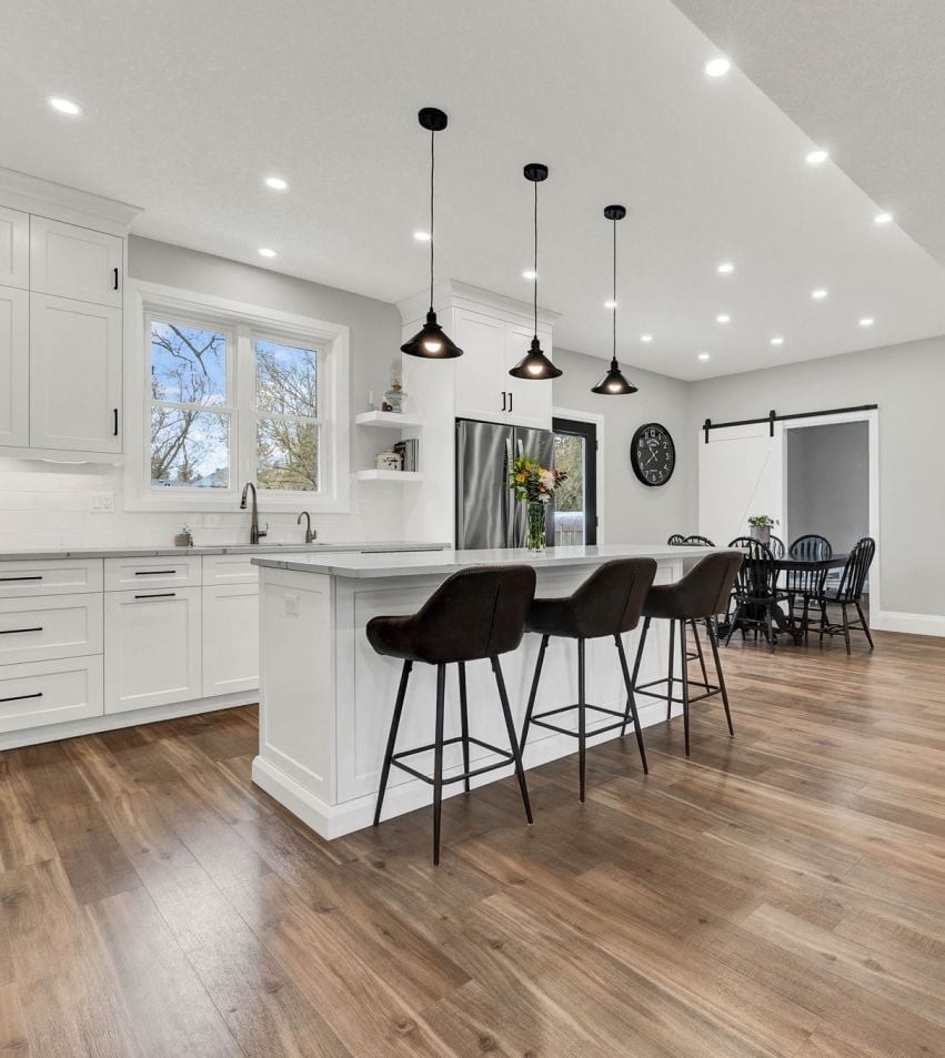 Bright white kitchen with a large island, dark bar stools, pendant lights, and warm wood flooring.