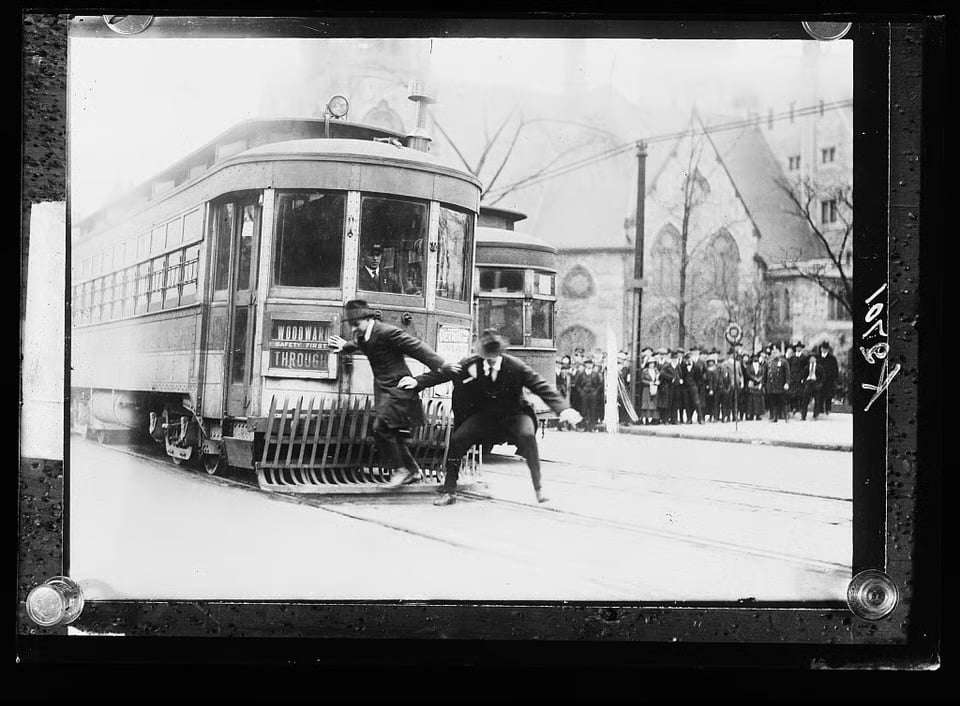 Historic Woodward Avenue streetcars with two men beside the lead car and a crowd outside a stone building.
