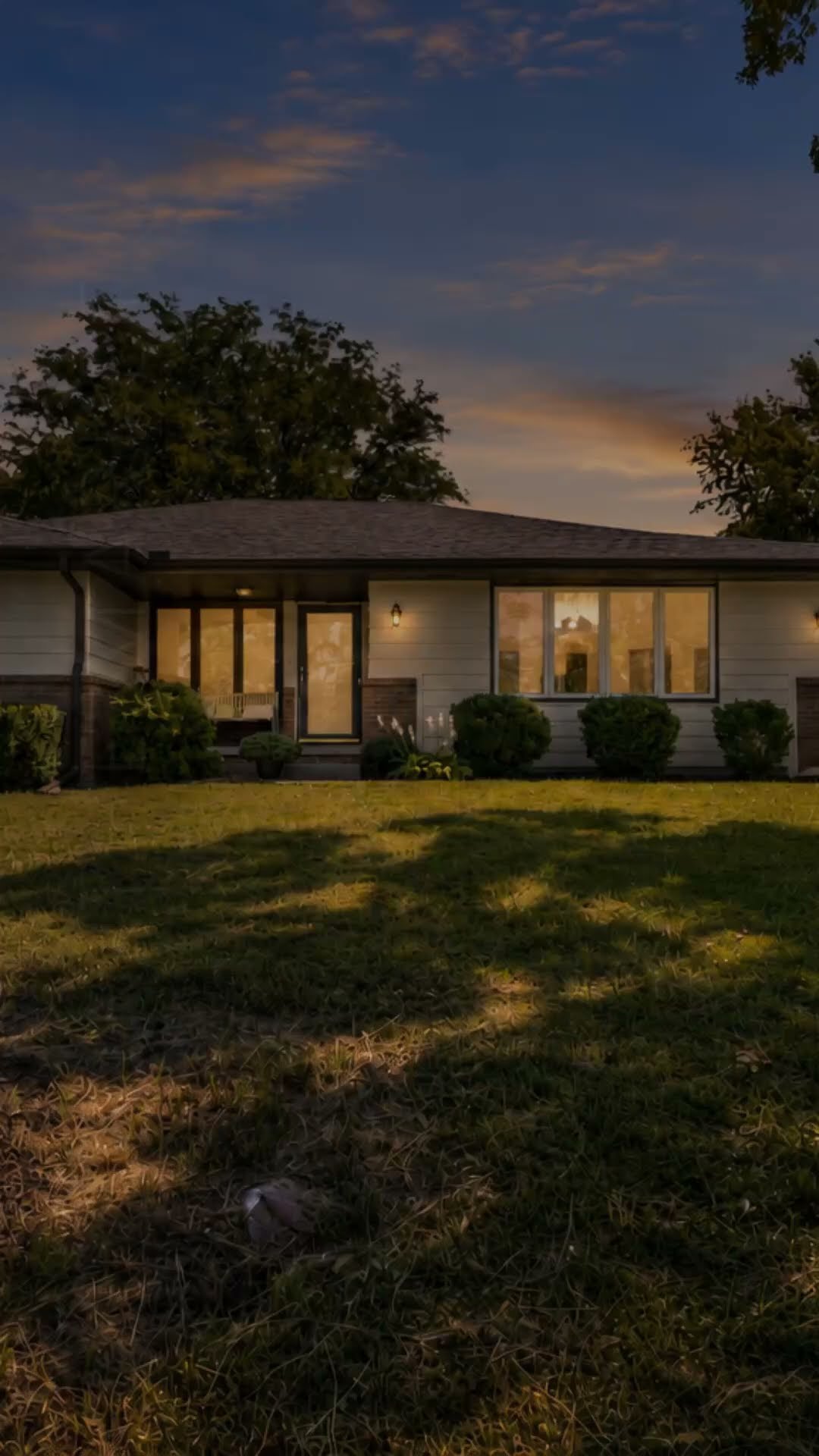 Gray ranch-style home at dusk with warmly lit windows, trimmed shrubs, and a broad lawn.