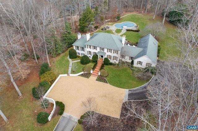 Aerial view of a white house with a circular pool and expansive paved driveway among leafless trees.