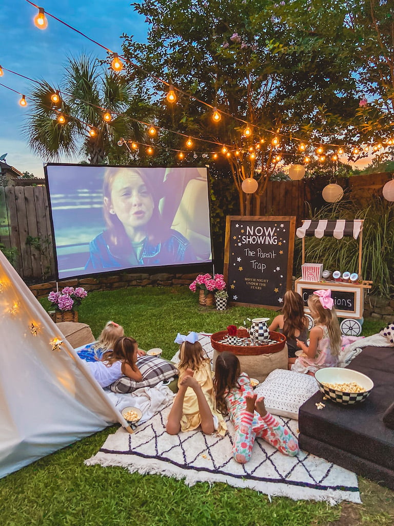 Children watch The Parent Trap outdoors beneath string lights, with blankets, popcorn, and a concession cart.