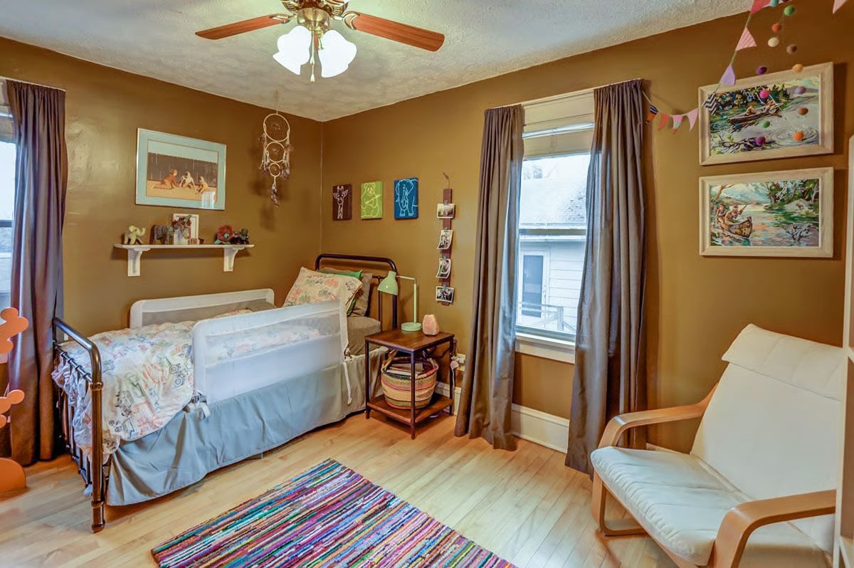 Bedroom with two single beds, warm brown walls, framed artwork, and a colorful striped rug.