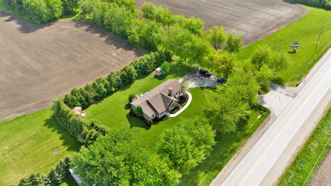 Aerial view of a rural home with expansive lawns, mature trees, and fields beside the road.