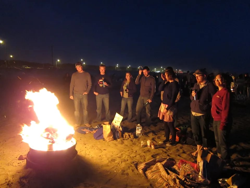 people gathered around a beach bonfire at Ocean Beach during sunset, with the sun setting over the Pacific Ocean and the sky painted in vibrant colors