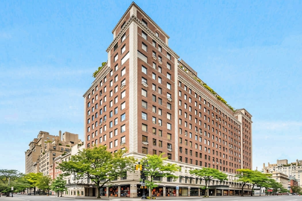 Red-brick corner apartment building with pale stone trim, storefronts, and leafy street trees.