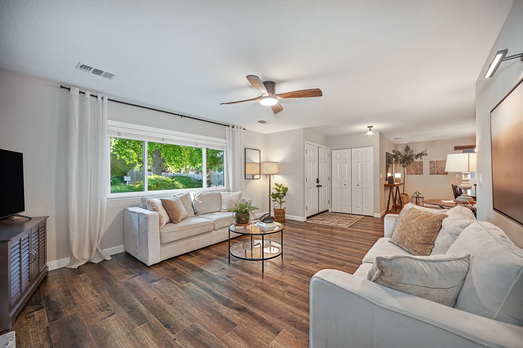 Living room with two light sofas, a round glass coffee table, and a large window overlooking greenery.