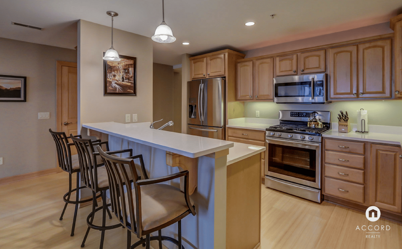 Condo kitchen with a white island, bar stools, stainless steel appliances, and maple cabinets.