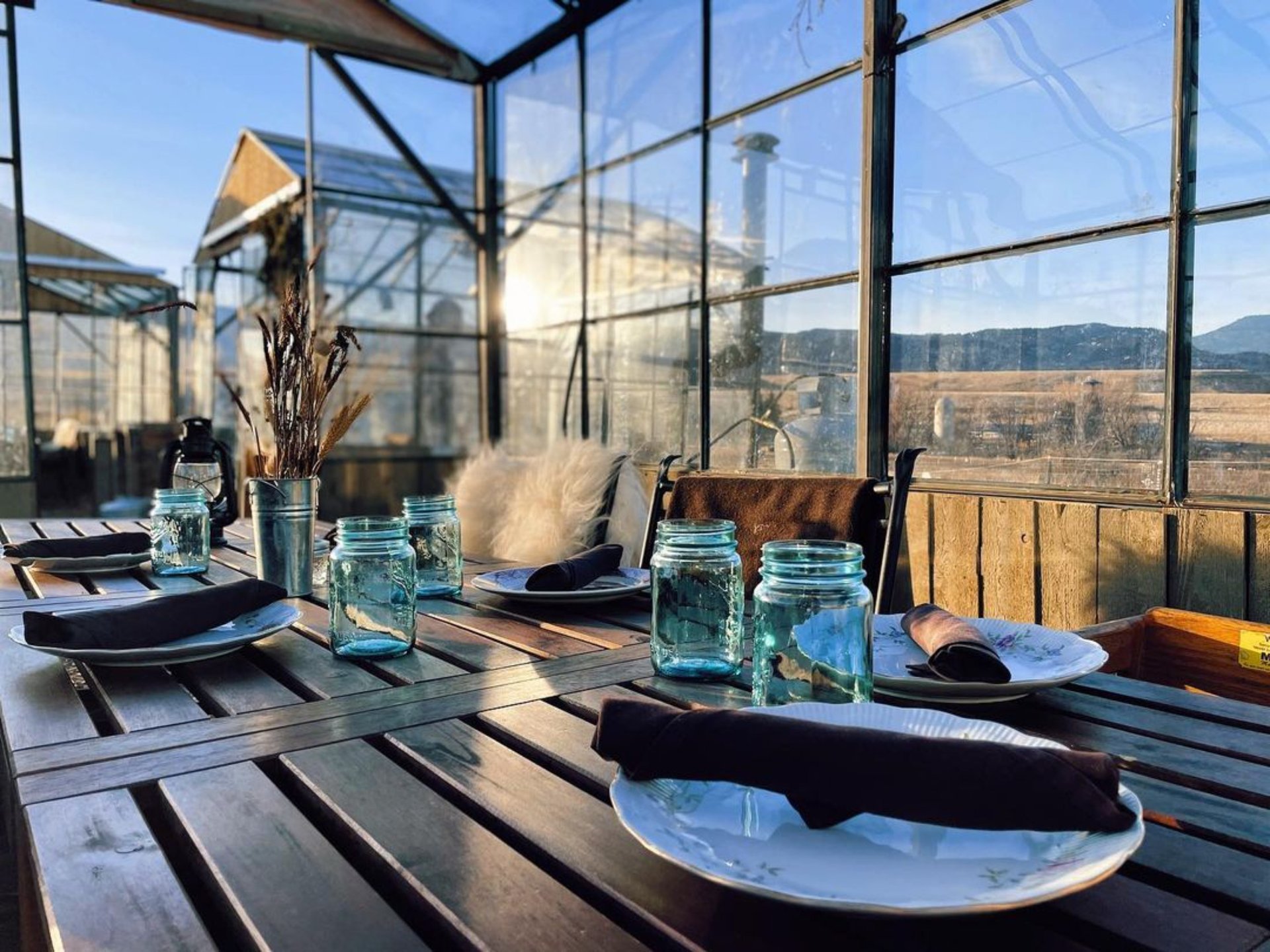 Restaurant patio table set with plates and blue glass jars, overlooking golden fields and mountains.