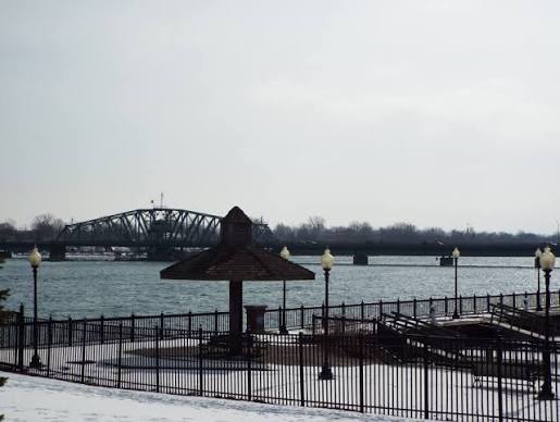 Snowy waterfront park with a fenced promenade, gazebo, and steel bridge across the river.