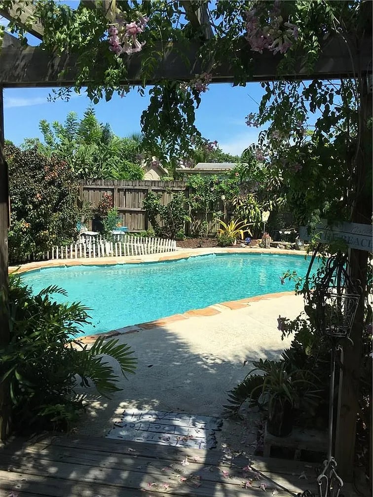 Backyard swimming pool framed by flowering vines, tropical plants, and a wooden privacy fence.