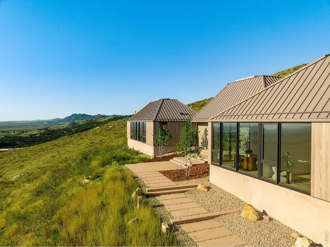 Modern home on a grassy mountainside, with angular metal roofs, large windows, and mountain views.