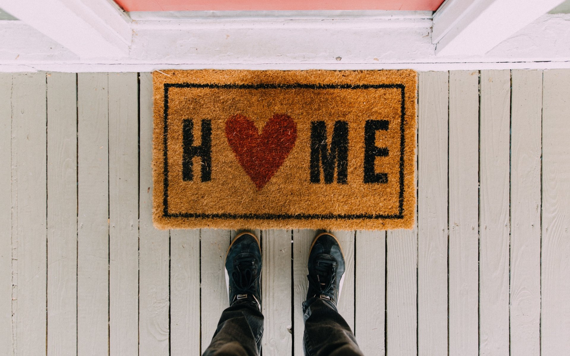 A visitor stands at a porch doormat printed with “H ♥ ME.”