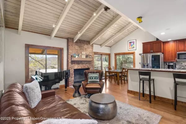 Living room with vaulted beam ceiling, stone fireplace, grand piano, and open kitchen.