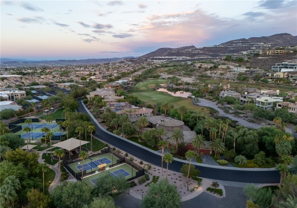 Aerial view of MacDonald Highlands in Henderson at sunset with tennis and pickleball courts, palm-lined estates, a golf hole with a pond and the McCullough Range