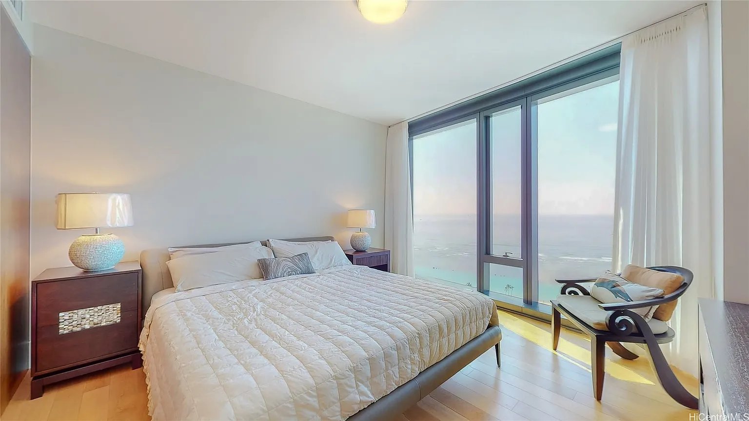 Furnished bedroom with floor-to-ceiling ocean-view windows and a chair beside the bed.
