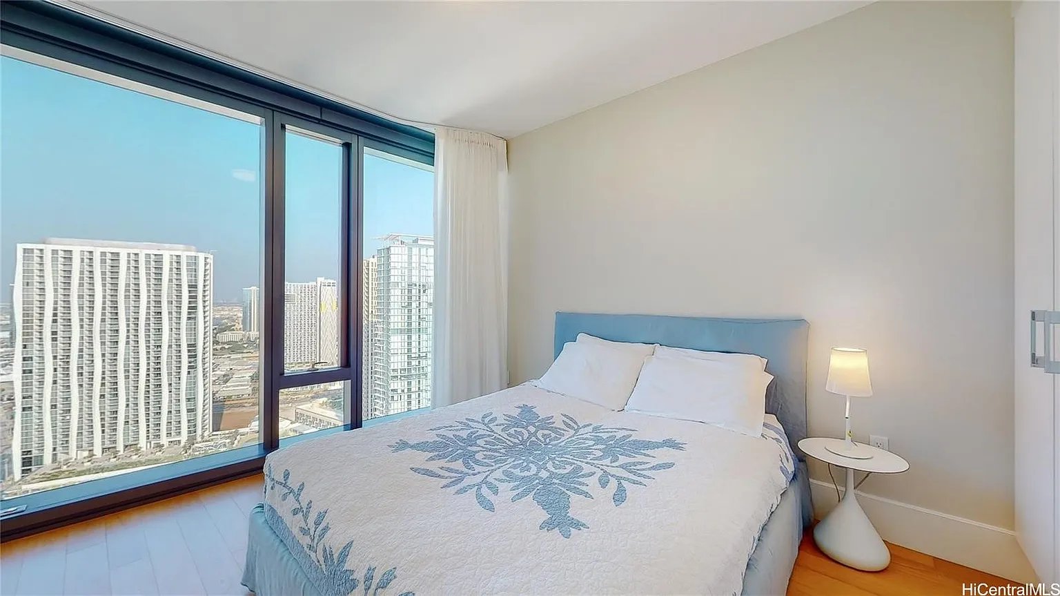 High-rise bedroom with a blue patterned quilt and floor-to-ceiling windows overlooking city towers.