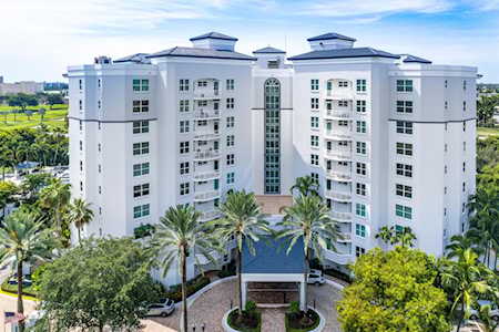 White condominium building with balconies, a central glass facade, and palm-lined entrance.
