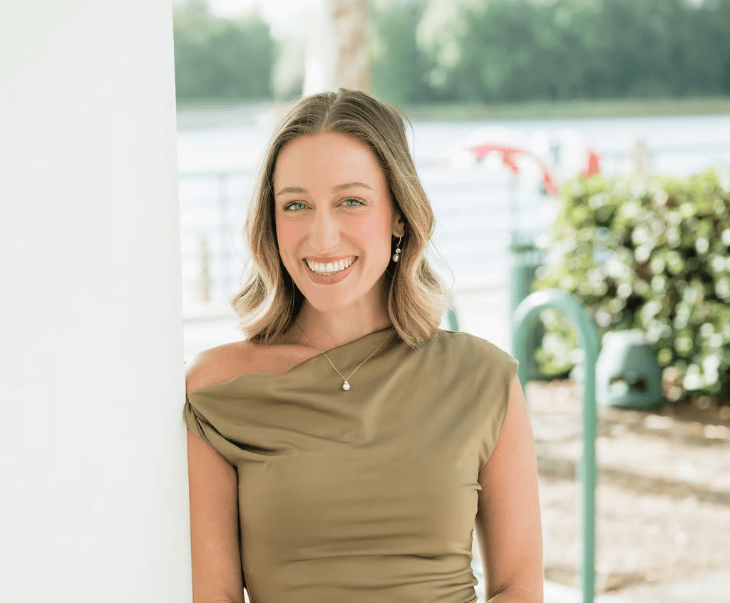 Smiling woman in an olive draped-neck top, photographed beside a waterfront patio.
