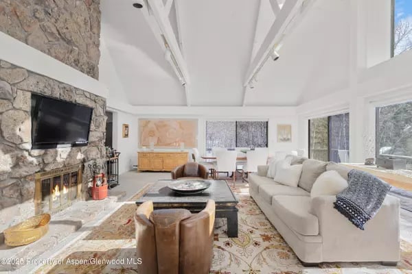 Living room with a tall stone fireplace, vaulted ceiling, and large windows overlooking snowy trees.