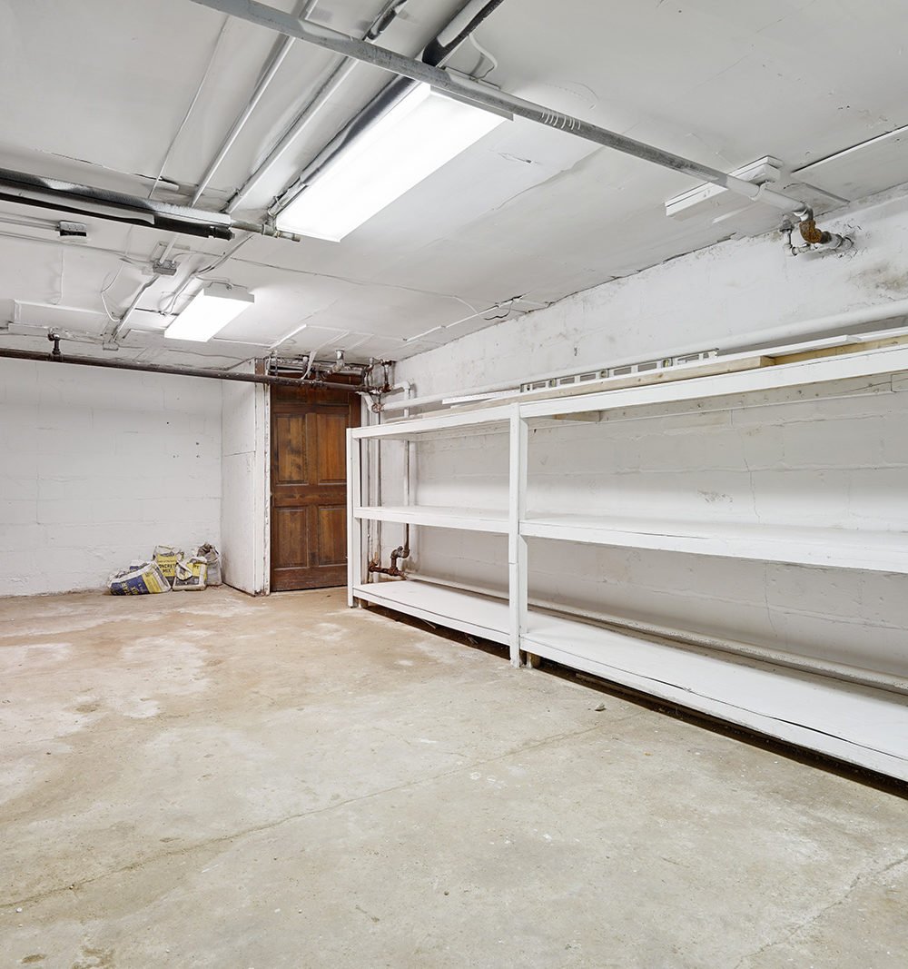 Basement storage room with long white shelves, exposed pipes, and a wooden door.