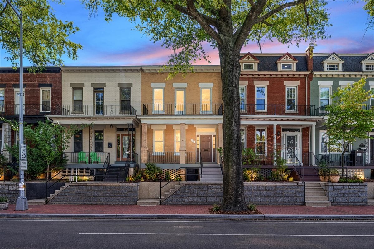 Tan-brick rowhouse with a raised stoop and balcony, framed by a large street tree.