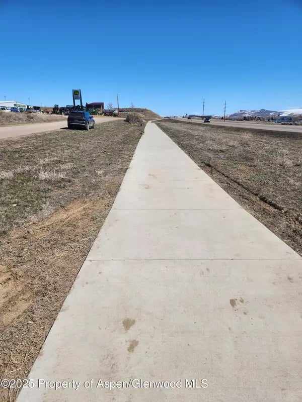 Concrete sidewalk through dry grass beside a road, with distant snow-dusted mountains.