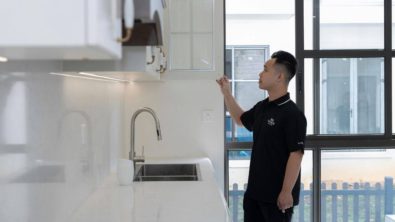 A person in a black shirt stands in a modern kitchen, examining a glass-fronted cabinet. The space is bright with sleek countertops and a large window.