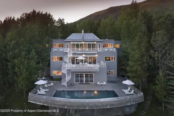 Modern multi-level home surrounded by forest, with expansive decks and an illuminated pool.