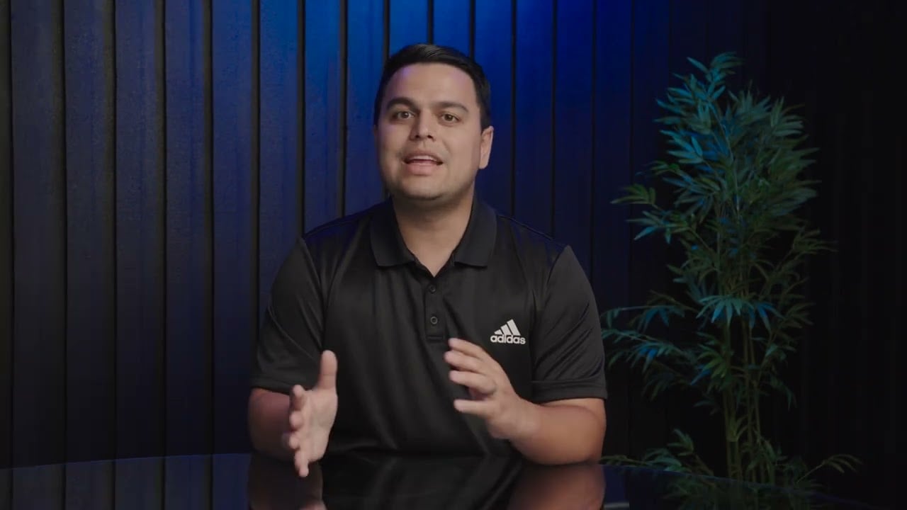 Man speaking to camera in a black Adidas polo against a blue-paneled backdrop.